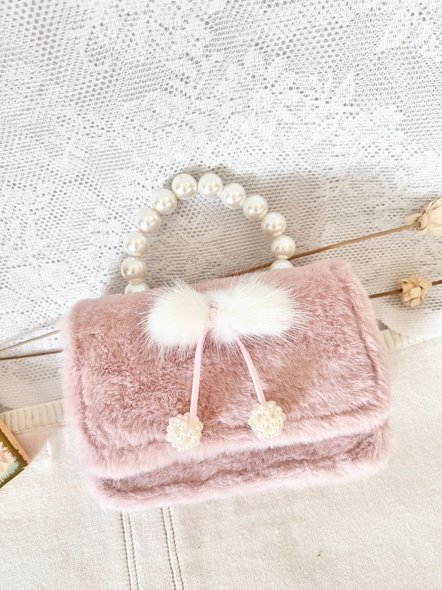 A small blush pink faux fur handbag with a pearl-bead handle, a fluffy white pom-pom, and two pearl-topped drawstrings, styled on a white lace fabric background.