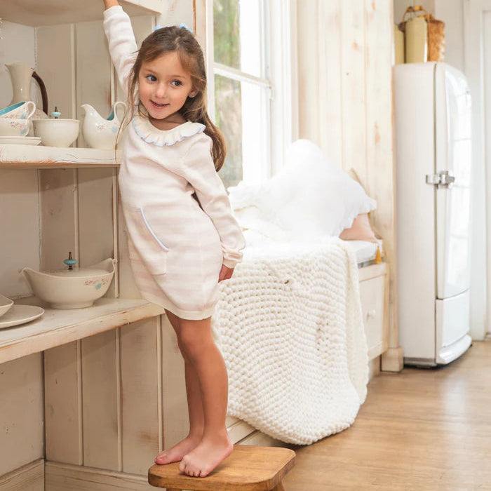 Young girl in a white dress standing on a wooden stool in a kitchen.