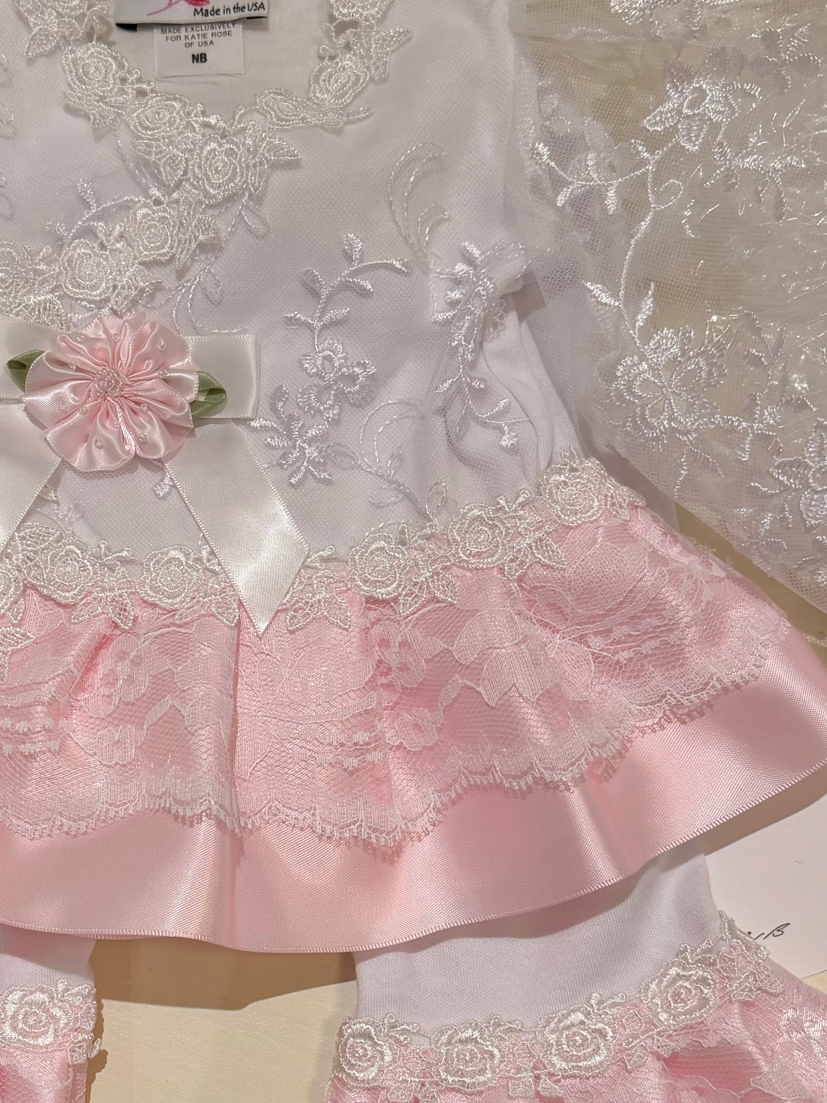 A close-up of a baby girl's two-piece outfit featuring a white long-sleeve top with intricate white floral embroidery, a white lace collar, a pink satin ruffled hem, a matching pink satin bow, and a pink fabric flower applique.