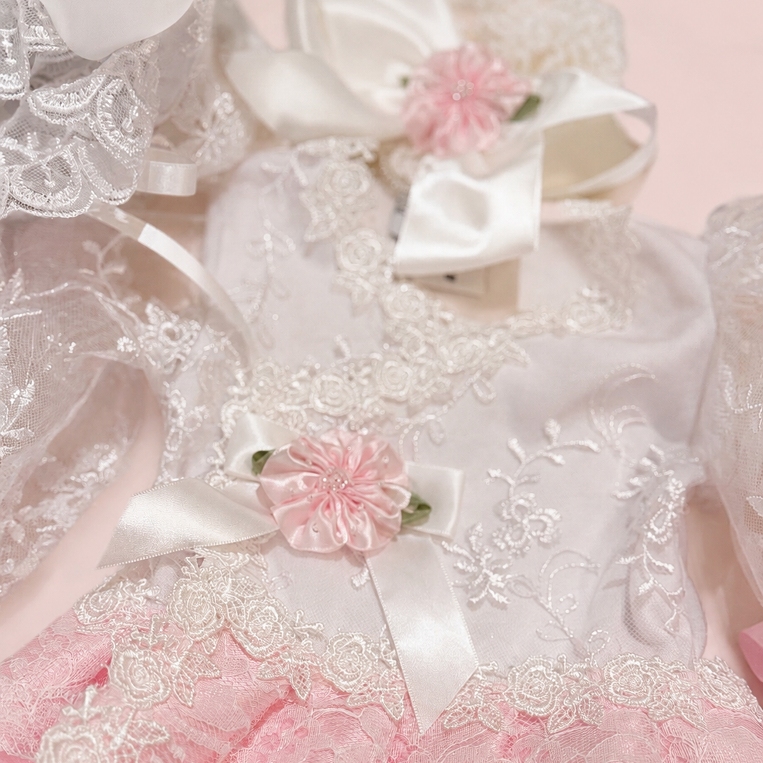 A close-up of a delicate baby dress featuring a white satin bodice with intricate floral embroidery, a pink lace ruffled hem, and matching pink fabric accents, adorned with white and pink satin bows and fabric flowers.