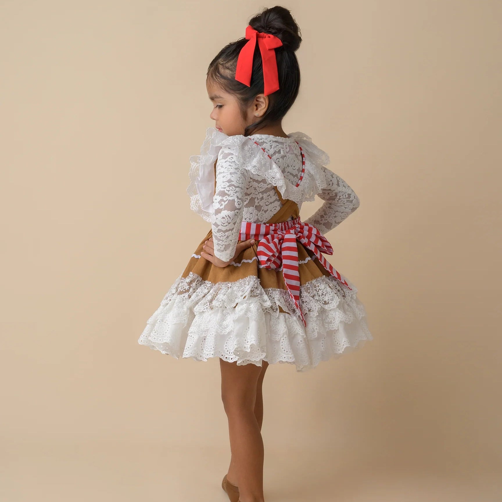 Young girl in a white lace dress with a red bow standing against a beige background