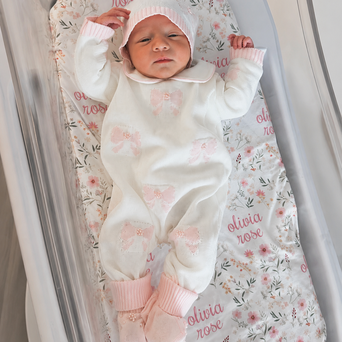 Newborn baby in a white outfit with pink accents lying on a floral-patterned blanket.