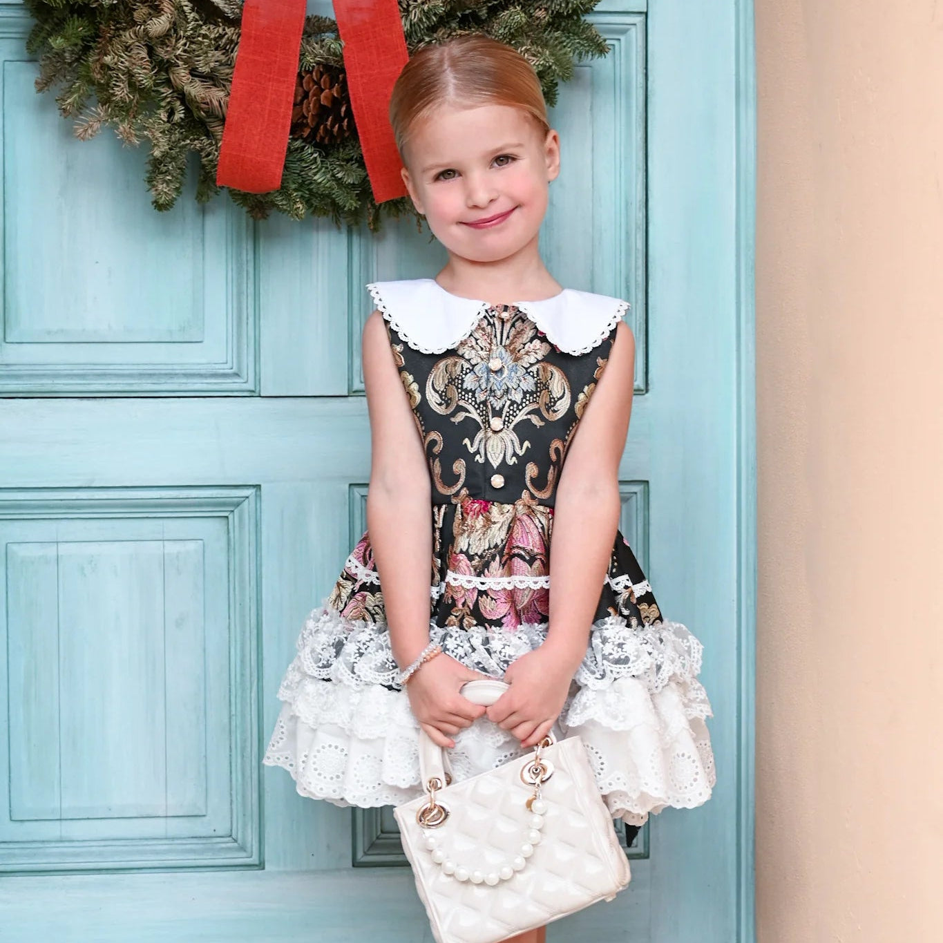 Young girl in a decorative dress standing in front of a blue door with a wreath.