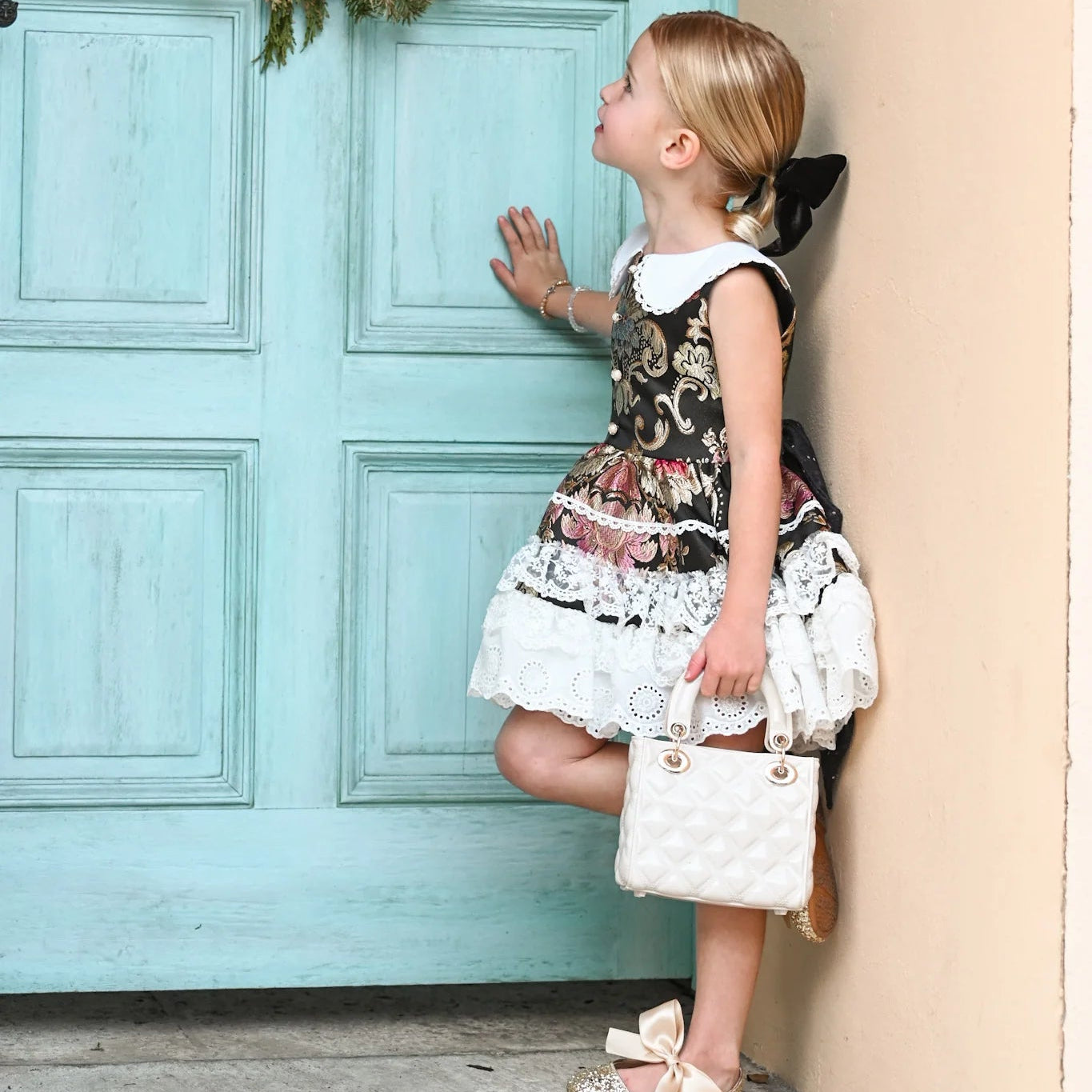 Young girl in a floral dress standing next to a turquoise door with a wreath.