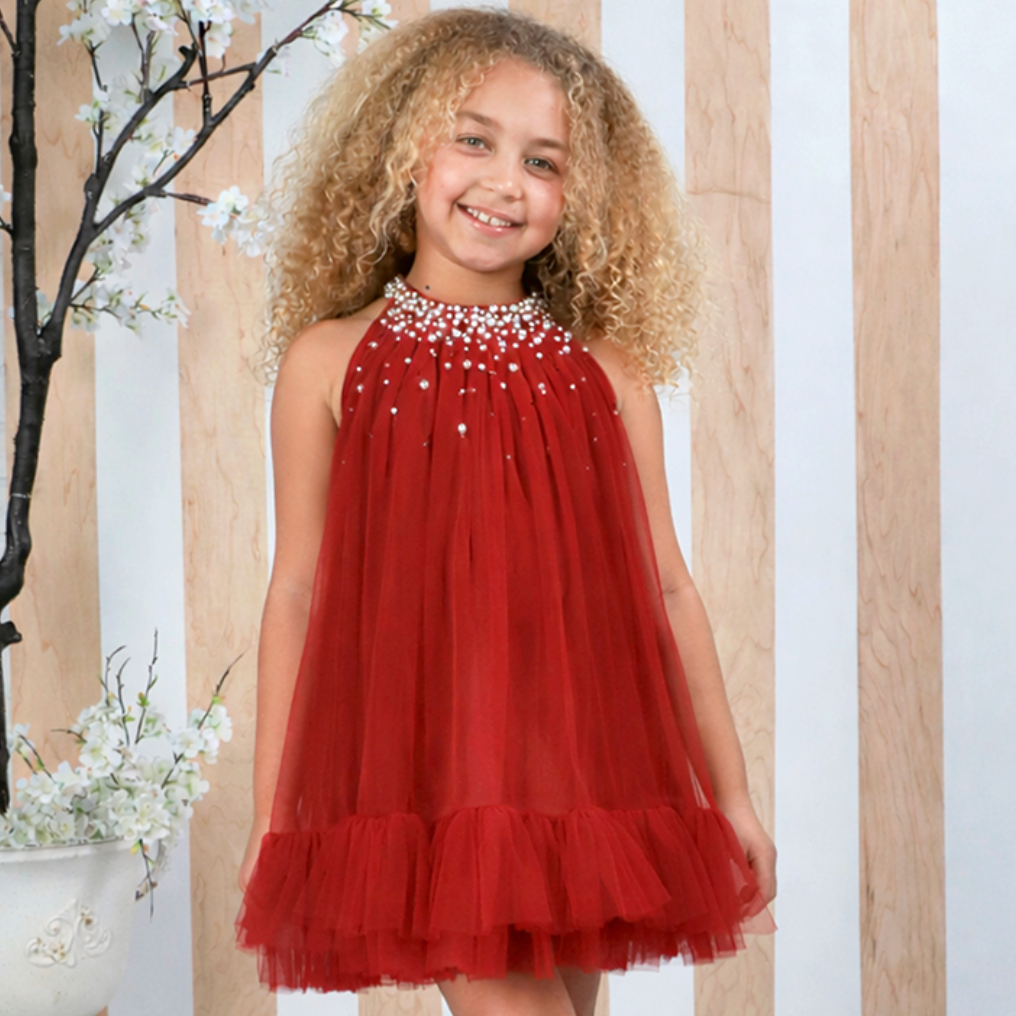 A young girl with curly blonde hair wearing a rust red sleeveless tulle dress with a beaded halter neckline and white lace-up boots, standing in front of a white and light wood paneled wall.